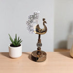 Decorative peacock sculpture on a wooden surface with a potted plant in the background