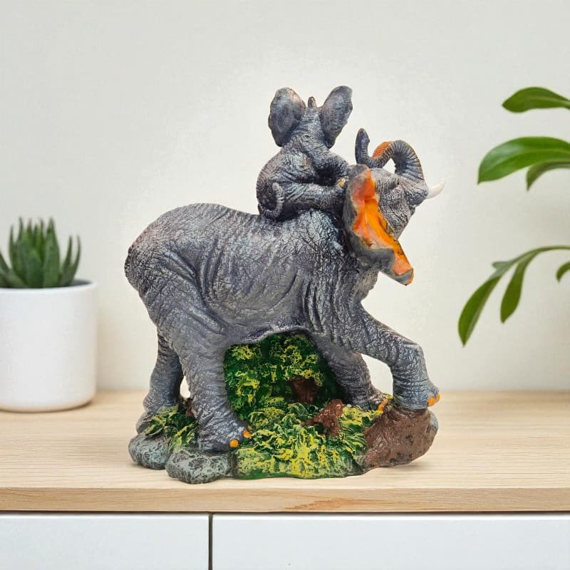 Decorative figurine of an elephant with a smaller animal on its back, placed on a wooden surface with plants in the background.