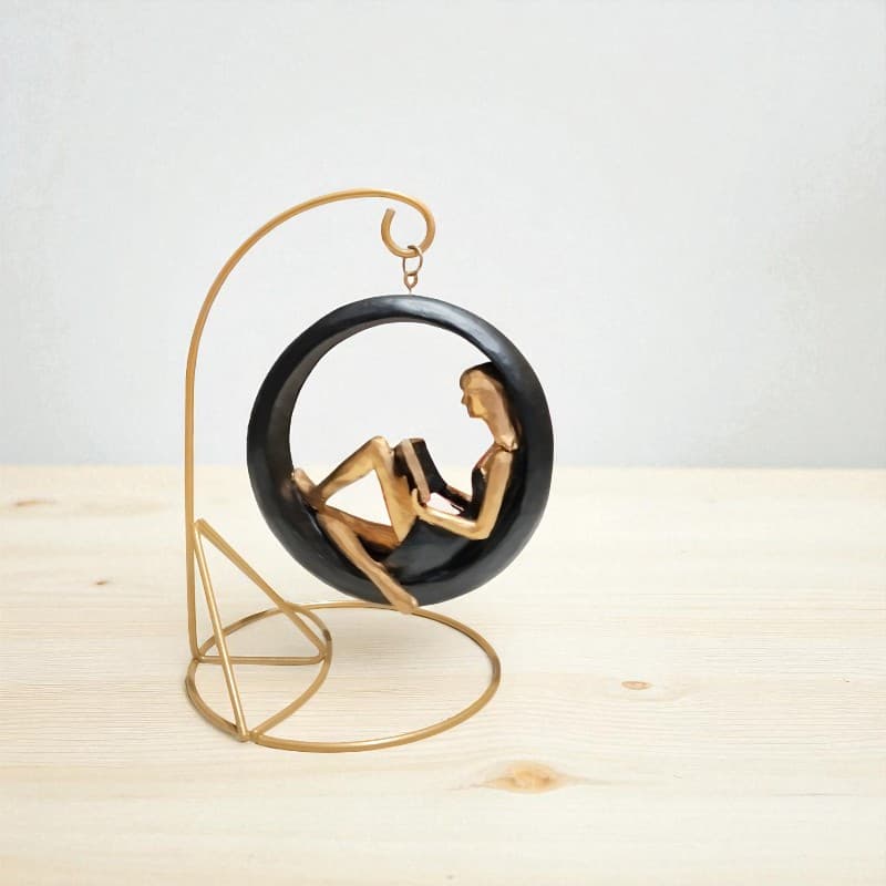 Decorative sculpture of a woman reading a book in a black chair with a gold stand on a light wooden surface.