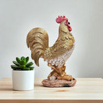 Decorative rooster statue next to a small potted plant on a wooden surface with a light gray background