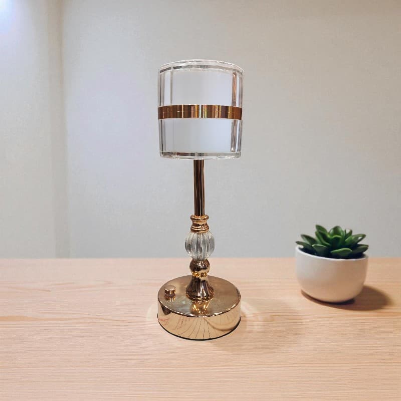 Small table lamp with gold accents on a wooden surface next to a potted plant.