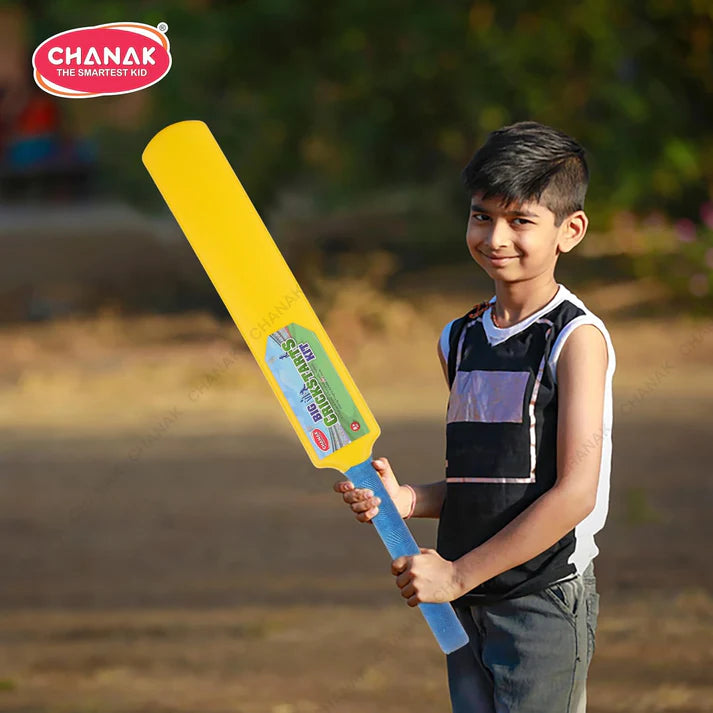 Child holding a large yellow and blue cricket bat outdoors with 'CHANAK' branding.