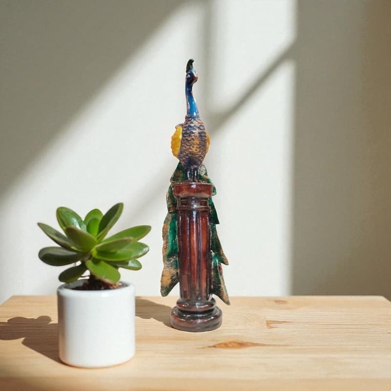 Decorative peacock figurine next to a potted plant on a wooden surface with a neutral background
