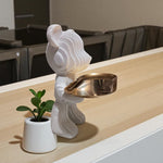 Decorative white sculpture with a gold bowl on a wooden surface, next to a small potted plant.