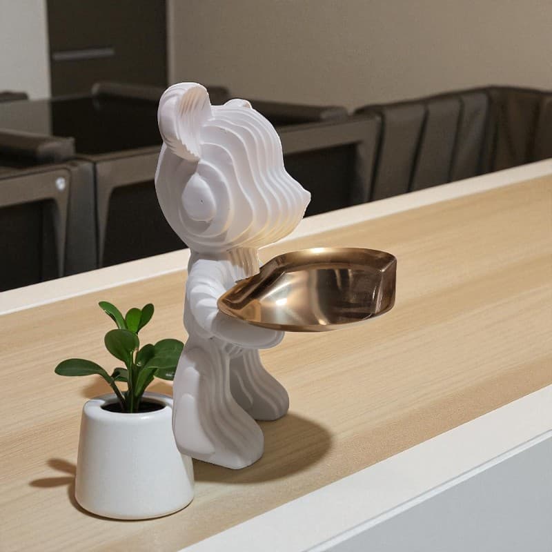 Decorative white sculpture with a gold bowl on a wooden surface, next to a small potted plant.