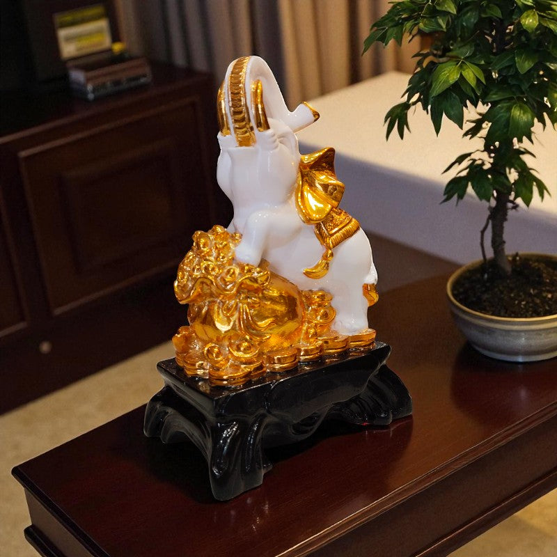 Decorative figurine of a white and gold elephant on a dark stand with a plant in the background.