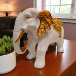 Decorative white elephant with gold accents on a wooden surface next to a potted plant.