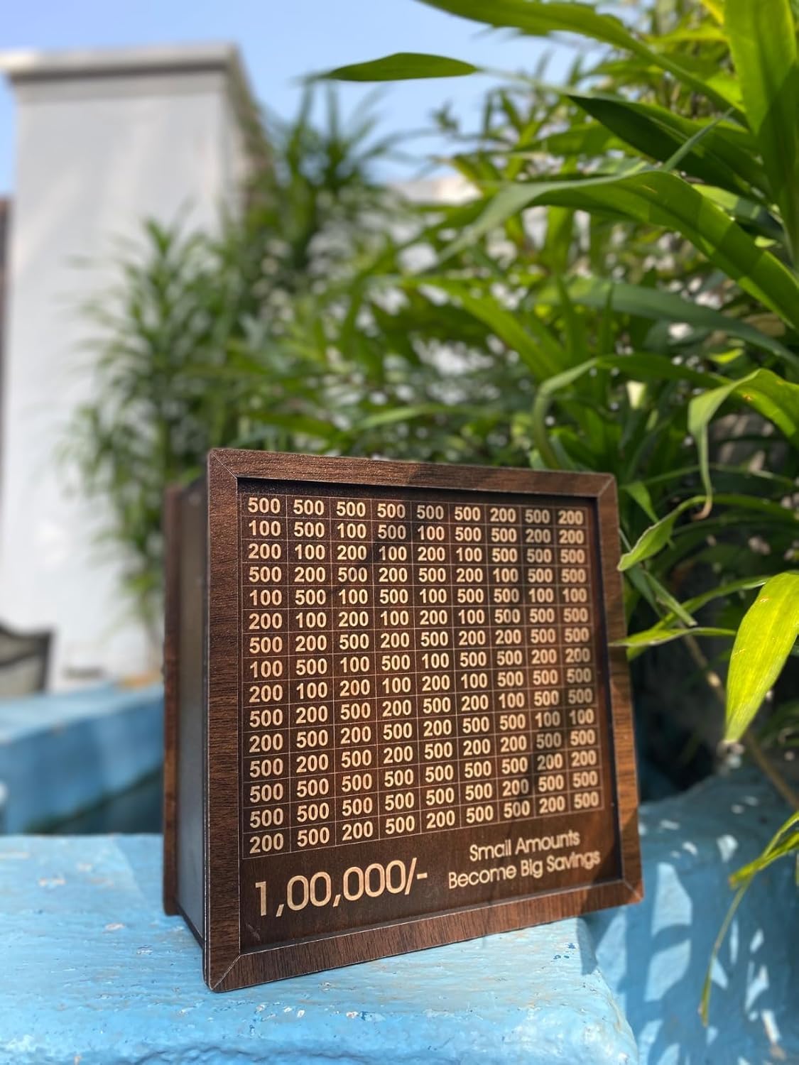 Front view of wooden piggy bank with 1 lakh savings grid placed outdoors with green plants