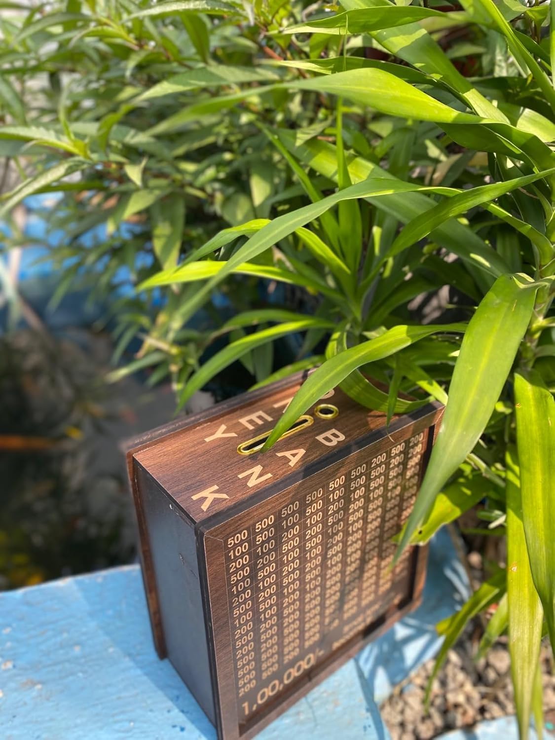 Side and top view of wooden 1 lakh piggy bank showing coin slot and engraved alphabets